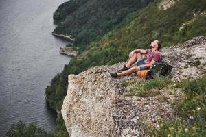 Traveller with backpack sitting on top of mountain enjoying view above the water surface. Traveling along mountains and coast, freedom and active lifestyle concept photo