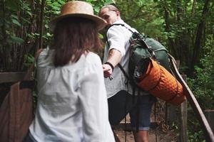 Two hikers with backpacks on the back in nature. Man and woman holding hands while walk on a summer day