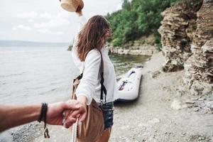 Portrait of happy young woman in hat holding man by hand and going to the boat on the lake in the mountains photo