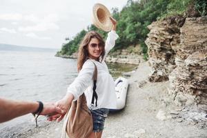 Portrait of happy young woman in hat holding man by hand and going to the boat on the lake in the mountains photo