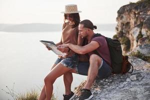 Hugging couple with backpack sitting on top of rock mountain enjoying view coast a river or lake. Traveling along mountains and coast, freedom and active lifestyle concept photo