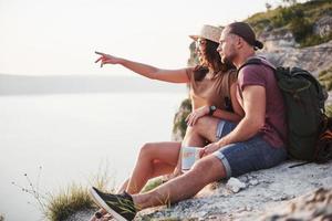 Hugging couple with backpack sitting on top of rock mountain enjoying view coast a river or lake. Traveling along mountains and coast, freedom and active lifestyle concept