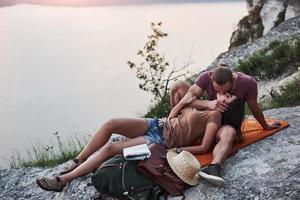 Hugging couple with backpack sitting on top of rock mountain enjoying view coast a river or lake. Traveling along mountains and coast, freedom and active lifestyle concept photo