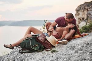 Hugging couple with backpack sitting on top of rock mountain enjoying view coast a river or lake. Traveling along mountains and coast, freedom and active lifestyle concept photo