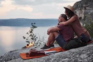 Hugging couple with backpack sitting near the fire on top of mountain enjoying view coast a river or lake. Traveling along mountains and coast, freedom and active lifestyle concept
