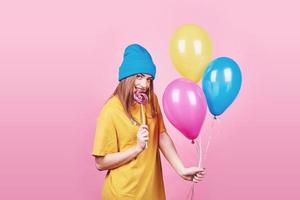 Cute funny girl in blue cap portrait holds an air colorful balloons and lollipop smiling on pink background. Beautiful multicultural Caucasian girl smiling happy photo