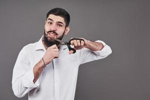 No more beard. Portrait of handsome young man cutting his beard with scissors and looking at camera while standing against grey background. New trend photo