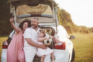 bastante joven matrimonio y su hija descansan en la naturaleza. la madre, el padre y la niña están sentados en el maletero del coche abierto foto