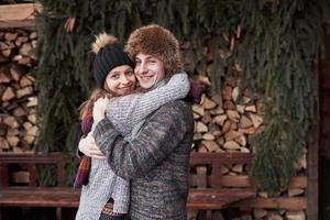 christmas and couple concept - smiling man and woman in hats and scarf hugging over wooden country house and snow background