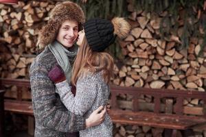 christmas and couple concept - smiling man and woman in hats and scarf hugging over wooden country house and snow background