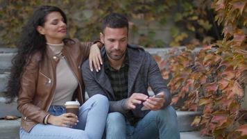 Handsome young couple sitting on outdoor stairs and using mobile phone on a autumn day