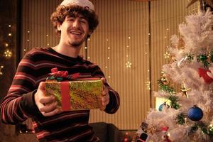 A portrait of a caucasian young man happiness smiling, looking at the camera, and handing out presents during a New Year's Eve party, in a home decorated with white Christmas trees and ornaments.