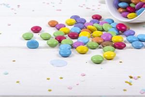 Bombones de caramelo de colores esparcidos sobre fondo de tablero de madera blanca foto