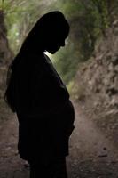 Silhouette of a pregnant woman touching and looking at her belly. Outdoor portrait, nature background. Figure against the light. . photo