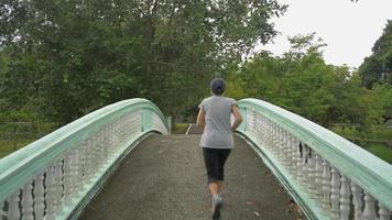 Asian middle aged woman jogging on the old bridge over a natural pond in a public park. video