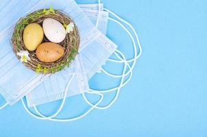 Nest with brightly dyed eggs with medical masks on blue background, space for text. Easter holiday during the Coronavirus pandemic photo