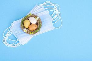Nest with brightly dyed eggs with medical masks on blue background, space for text. Easter holiday during the Coronavirus pandemic photo