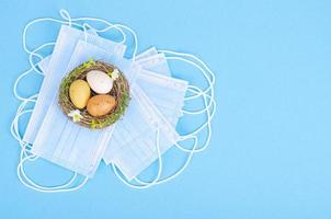 Nest with brightly dyed eggs with medical masks on blue background, space for text. Easter holiday during the Coronavirus pandemic photo