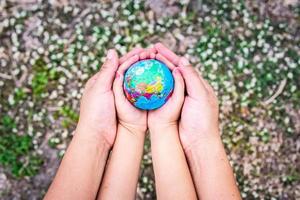 Closeup mother hand embraces daughter hand embracing our globe in our palms on grounded background with green grass and white flowers. Blur background. Concept to help take care of our world. photo