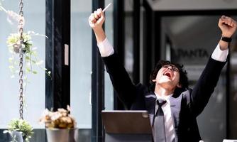 El hombre emocionado y lleno de alegría recibió buenas noticias en una carta, promoción laboral o un gran resultado de examen, feliz hombre de negocios o estudiante celebra el éxito, gritando, levantando las manos, sentado en el lugar de trabajo con una computadora portátil foto