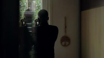 Hombre de pie y llamando con smartphone mientras mira a través de la ventana