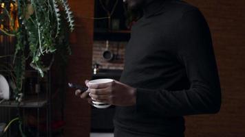 Mann am Fenster, der auf dem Smartphone tippt und die Tasse hält, dreht sich zum Kameraobjektiv