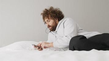 Hombre acostado en la cama con smartphone mientras mueve la cabeza y tararea