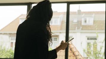 Woman standing next to window talking and scrolling on smartphone video
