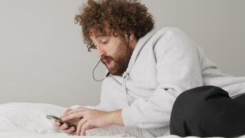 Man lying on bed busy with smartphone whilst moving head and humming video