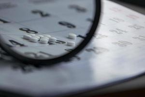 Conceptual photo with pills under a magnifying glass. White pills on a calendar background.