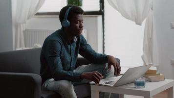 homme afro-américain souriant authentique utilisant un ordinateur portable avec des écouteurs, travaillant à domicile