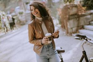 Young woman with mobile phone drink coffee to go by the bicycle on autumn day photo
