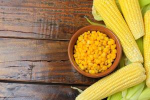 Raw corn kernels, inside ceramic bowl, next to corn on the cob on rustic wooden table. photo