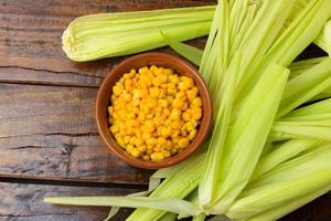 Raw corn kernels, inside ceramic bowl, next to corn on the cob on rustic wooden table. photo