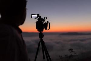 Cameraman and video camera operator working with his equipment shooting sunrise foggy landscape photo