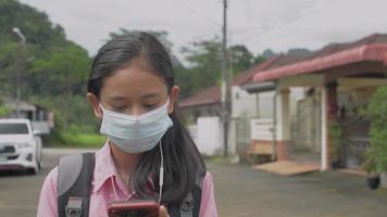 vrouwelijke tienerstudent die een gezichtsmasker draagt dat naar sociale video op smartphone kijkt terwijl hij naar huis loopt.