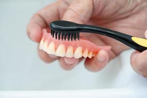 Asian senior or elderly old woman patient use toothbrush to clean partial denture of replacement teeth. photo