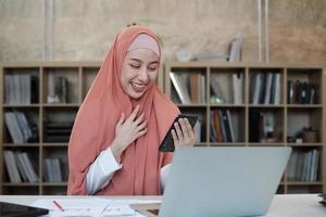 hermosa mujer de negocios que persona de sexo femenino islámico está hablando por la comunicación de su teléfono móvil y sonriendo feliz porque orden en línea de Internet en la computadora portátil, estilo de vida moderno, ocupación de comercio electrónico. foto