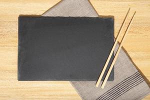 Flat lay of Blank Black slate Tray, chopsticks and linen napkin on wooden table. Template Mock up photo