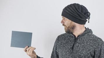 A Bearded Surprised Hipster Man On A White Background In A Knitted Hat Holds In His Hands A Blank Gray Carton Sign For Advertising. Copy space. High Quality Photo