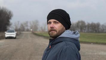 Portrait Of An Attractive Hipster Man With A Beard In A Knitted Hat. Man Against Blurry Car Background photo