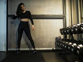 sporty brunette in black leggings and top is standing tall on a shelf with dumbbells in the gym against the background of the lattice photo