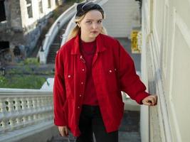 attractive blonde hipster girl in a knitted black hat and red jacket is standing on the stairs under the bridge photo