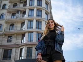 attractive blonde girl smiling in a denim jacket on the background of a modern building and blue sky photo