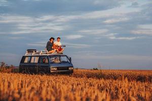 Couple man with a guitar and woman in a hat are sitting on roof of a car in a wheat field photo