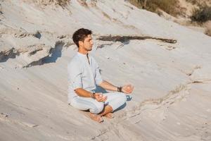 Man dressed in light clothing sits in meditation pose on sandy beach