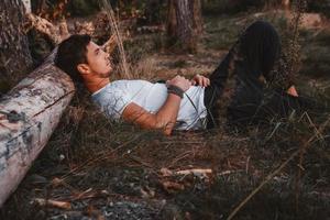 Man lies on grass, resting his head on a log in middle of forest relaxing