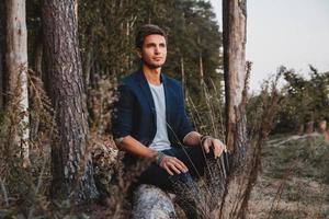 El hombre posando al aire libre en el bosque se sienta en un tronco vistiendo chaqueta marcada foto