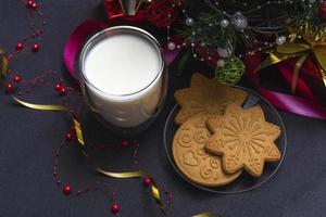 pan de jengibre y leche para santa. Composición navideña con galletas de jengibre y leche sobre un fondo negro con un árbol de navidad y un regalo.
