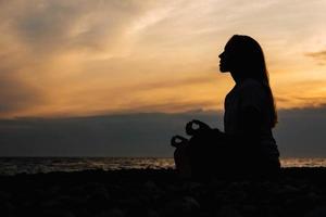 Silueta de una mujer en pose de meditación en la playa durante la puesta de sol surrealista en el fondo del mar y el cielo dramático